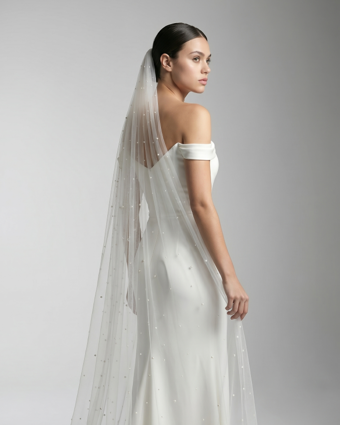 Eine Braut mit Bun, Perlenschleier und cleanen Kleid steht vor einer weiß-grauen Wand